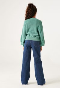 Zielony sweter wykonany na drutach o ryflowanej fakturze i luźnych rękawach w kształcie balonów, w połączeniu z szerokimi niebieskimi dżinsami o ciemnym kolorze.