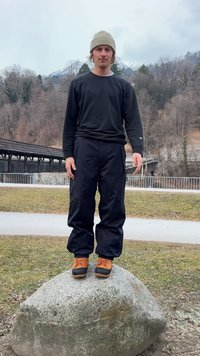Pantalon noir et chemise noire à manches longues portés par une personne debout sur une pierre. Chaussures marron-orange, bonnet gris clair ; arrière-plan en plein air.