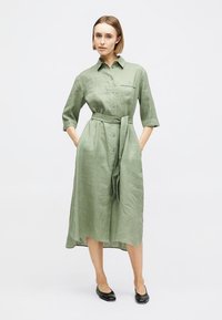 Femme debout, les mains dans les poches, portant une robe midi boutonnée vert clair avec une ceinture à la taille et des chaussures plates noires.