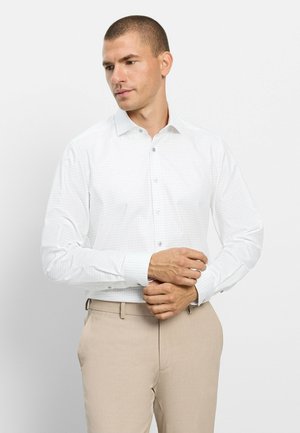 Uomo che sistema il polsino di una camicia bianca a maniche lunghe con un motivo a quadri discreto, indossa pantaloni beige su uno sfondo chiaro e uniforme.