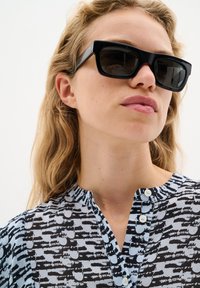 Blusa nera e azzurra chiara a fantasia con pattina abbottonata e colletto alla coreana, abbinata a occhiali da sole scuri e quadrati. Tessuto morbido.