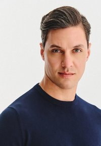 Portret osoby w granatowym swetrze z okrągłym dekoltem. Osoba ma krótkie, stylowe włosy i zielone oczy, a na twarzy neutralny wyraz.