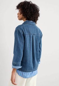 Dunkelblaue Denimjacke mit lockerer Passform, langen Ärmeln, Kragen und umgeschlagenen Manschetten, getragen über einem hellblauen gestreiften Hemd.