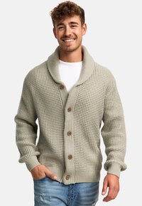 Cardigan de punto gris claro con cuello de chal, que presenta botones de madera, tejido texturizado y puños acanalados. Lleva puesto un T-shirt blanco.