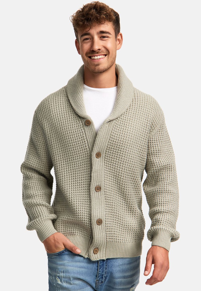 Cardigan de punto gris claro con cuello de chal, que presenta botones de madera, tejido texturizado y puños acanalados. Lleva puesto un T-shirt blanco.