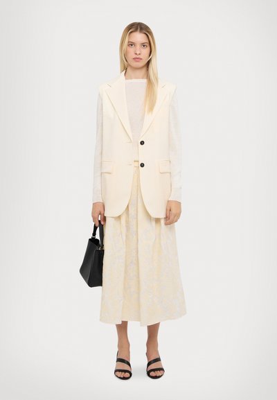 Cremefarvet ærmeløs blazer med to sorte knapper, parret med en lys gul nederdel med et diskret blomstermønster. Sort håndtaske og slidesandaler.