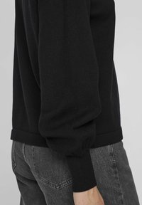 Pull en tricot noir avec un ourlet et des manches côtelés, affichant une coupe décontractée et une texture douce, associé à un pantalon en denim gris.