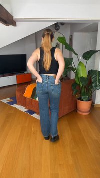 Kvinne med langt hår i hestehale, iført svart singlet og jeans, står med hendene i baklommene i en moderne stue.
