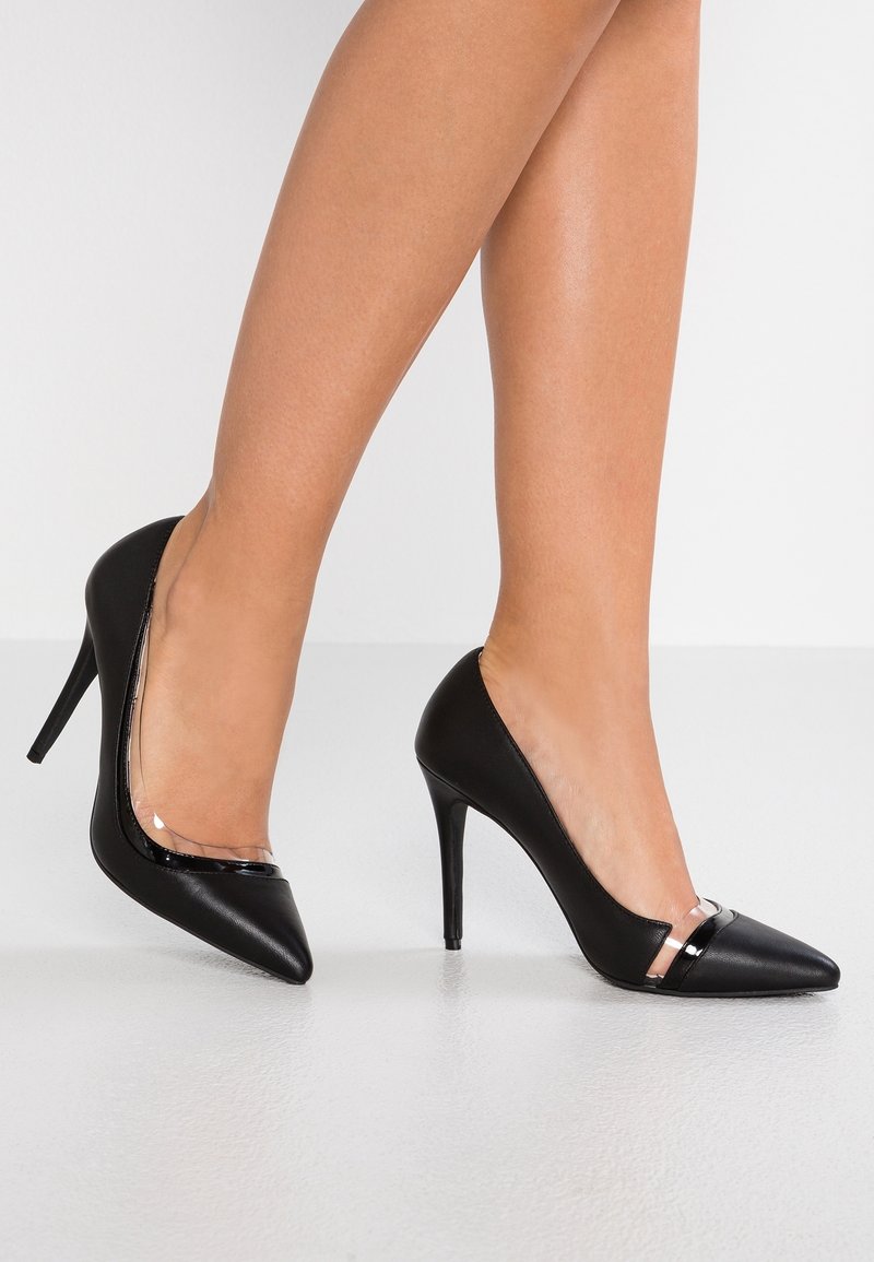 Femme portant des escarpins noirs à bout pointu avec des brides transparentes, posant avec un pied légèrement levé sur un fond uni.