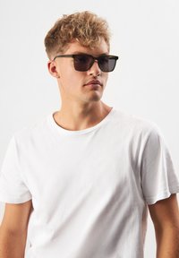 Svarta rektangulära solglasögon med mörka linser, placerade på en ljusgrön ram. Modellen bär en enfärgad vit t-shirt med korta ärmar.