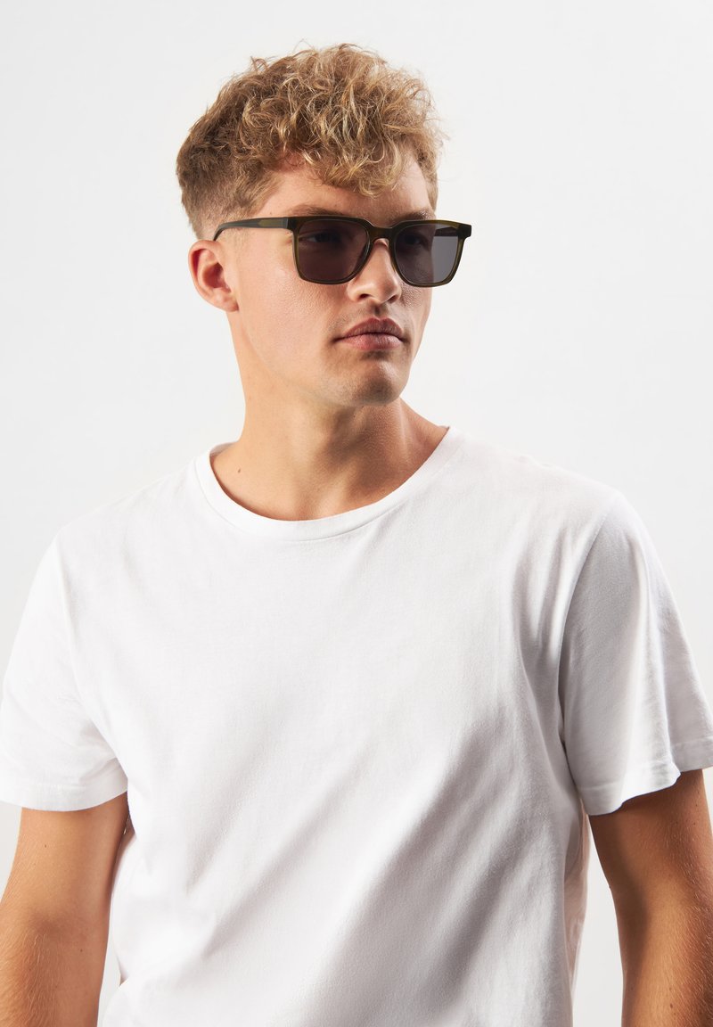 Svarta rektangulära solglasögon med mörka linser, placerade på en ljusgrön ram. Modellen bär en enfärgad vit t-shirt med korta ärmar.