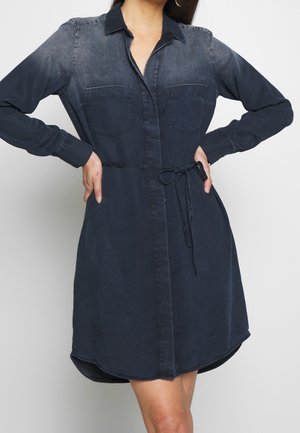 Femme portant une robe chemise bleu foncé à manches longues avec des poches avant et une ceinture nouée à la taille, posant avec les mains sur les hanches sur fond blanc.