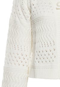Cardigan bianco lavorato a maglia con motivi testurizzati a zigzag e disegni a trama aperta. Lunghezza cropped con polsini e orlo a costine.
