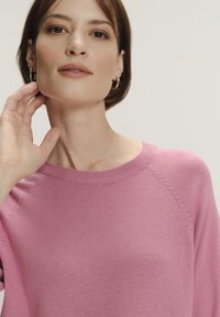 Jasnoróżowy sweter z okrągłym dekoltem, prążkowanym brzegiem i subtelnymi detalami teksturowymi na ramionach. Modelka dotyka szyi jedną ręką.