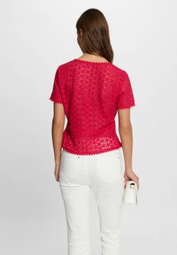 Haut en dentelle rouge à manches courtes et encolure festonnée, associé à un pantalon blanc. Le haut présente un design semi-transparent et texturé.