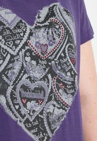 Fialová košile s velkým srdcovým grafickým vzorem, který obsahuje německý text a malé červené rhinestonové akcenty na několika srdcích.