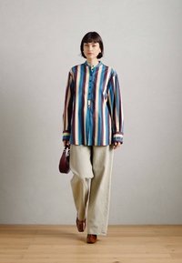 Ριγέ πουκάμισο σε μπλε, κόκκινο και κρέμ με κουμπωτό γιακά, συνδυασμένο με μπεζ παντελόνια σε φαρδιά γραμμή και καφέ παπούτσια, κρατώντας μια μπορντό τσάντα.