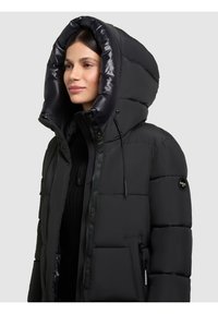 Sort pustejakke med oversize hætte, lynlås foran og knaplukning, med quiltet tekstur og sidelommer.