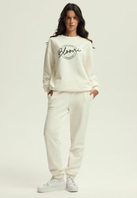 Cremfarbener Jogginganzug aus weichem Stoff, bestehend aus einem lässig geschnittenen Sweatshirt mit schwarzem Drucktext und passenden Jogginghosen mit elastischen Bündchen.