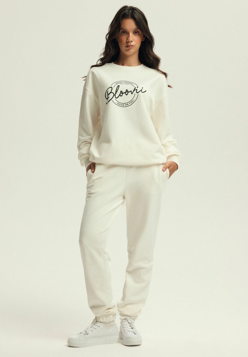 Cremfarbener Jogginganzug aus weichem Stoff, bestehend aus einem lässig geschnittenen Sweatshirt mit schwarzem Drucktext und passenden Jogginghosen mit elastischen Bündchen.