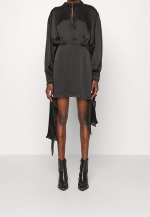 Kobieta ubrana w czarną satynową mini sukienkę z długimi rękawami o asymetrycznym kroju z marszczonymi mankietami i czarne botki, stojąca na białym tle.