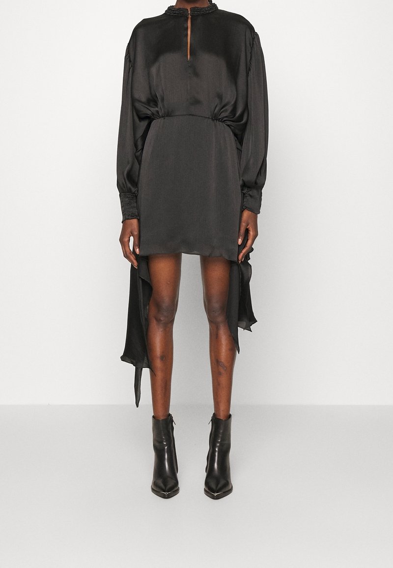 Femme portant une mini-robe noire en satin à manches longues avec ourlets froncés et bottines noires, debout sur un fond blanc.