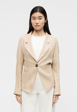 Νεαρή γυναίκα με ίσια σκούρα μαλλιά φοράει ένα μπεζ σακάκι με ένα κουμπί πάνω από λευκό πουκάμισο και λευκό παντελόνι, στέκεται μπροστά σε απλό φόντο.