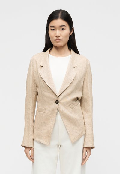 Jeune femme aux cheveux bruns lisses portant un blazer beige à un bouton sur une chemise blanche et un pantalon blanc, debout devant un fond uni.