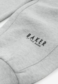 A szürke melegítőnadrág texturált anyagból készült, bordás mandzsettákkal és egy feltűnő fekete nyomtatott logóval rendelkezik, amelyen a "BAKER" és a "by TED BAKER" felirat szerepel az oldalon.