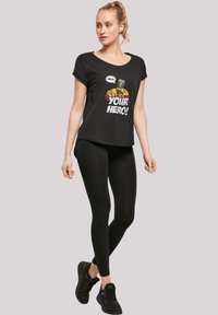 Black short-sleeve t-shirt featuring colorful graphic with text “YOUR HERO!” paired with black leggings and black sneakers.
