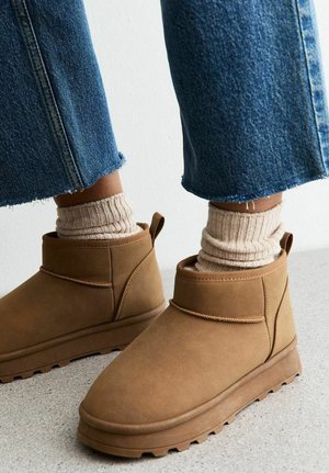 Botas de tobillo de ante marrón con un caña corta, suela de goma texturizada y un acento cosido. Combinadas con calcetines beige acanalados y jeans.