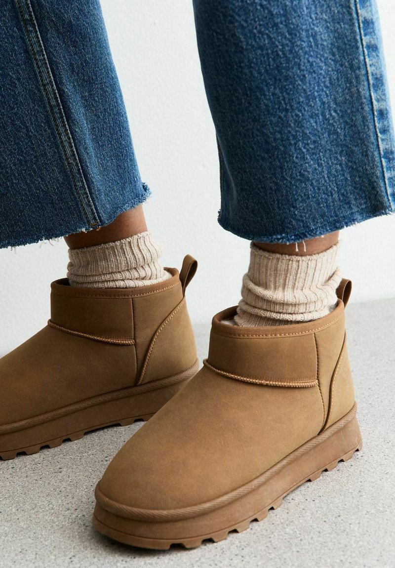 Botas de tobillo de ante marrón con un caña corta, suela de goma texturizada y un acento cosido. Combinadas con calcetines beige acanalados y jeans.