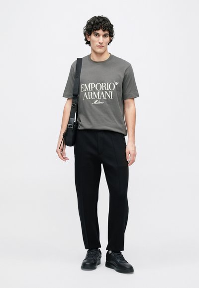 Siv�é bavlnené tričko s potlačou „EMPORIO ARMANI Milano“, skombinované s čiernymi nohavicami na mieru a čiernymi teniskami. Jednoduchá kabelka cez rameno ako doplnok.