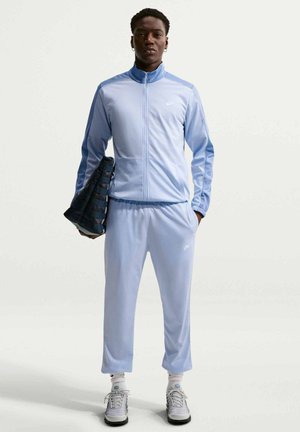 Osoba stojąca w jasnoniebieskim dresie Nike, białe trampki, trzyma czarną teksturowaną torbę w lewej ręce, zwrócona przodem.