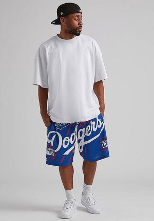 Uomo che indossa una t-shirt bianca oversize, pantaloncini blu dei Dodgers con dettagli rossi e bianchi, calzini bianchi, scarpe da ginnastica bianche e un cappellino nero indossato al contrario.