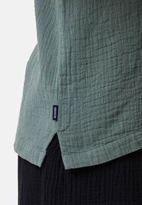 Chemise texturée vert clair à coupe décontractée, avec une fente sur le côté et une petite étiquette bleue portant la mention "SEASIDE" sur l'ourlet.