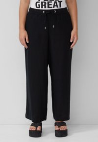 Schwarze weite Hosen aus weichem Stoff mit elastischem Bund und Kordelzug. Kombiniert mit schwarzen Plateausandalen, die die Zehen zeigen.