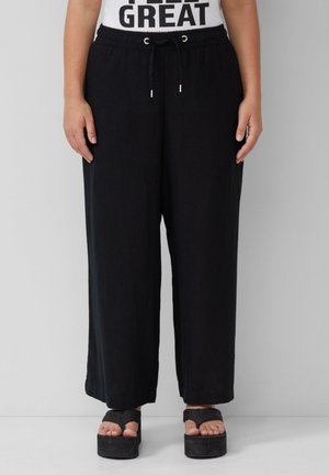 Schwarze weite Hosen aus weichem Stoff mit elastischem Bund und Kordelzug. Kombiniert mit schwarzen Plateausandalen, die die Zehen zeigen.