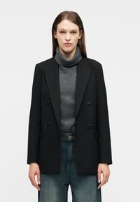 Γυναίκα που φοράει μαύρο σακάκι με διπλό πέτο πάνω από γκρι πουλόβερ με ζιβάγκο και μπλε τζιν, στέκεται μπροστά σε απλό λευκό φόντο.