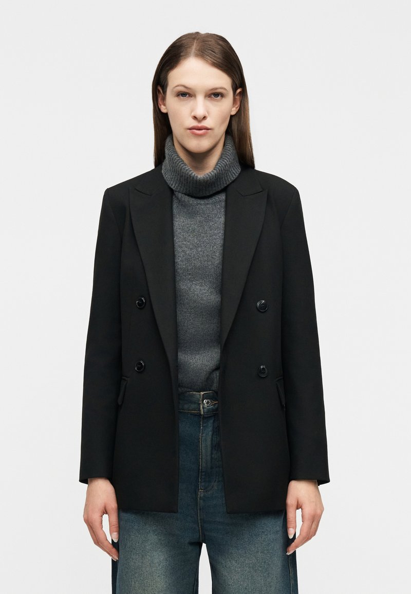 Γυναίκα που φοράει μαύρο σακάκι με διπλό πέτο πάνω από γκρι πουλόβερ με ζιβάγκο και μπλε τζιν, στέκεται μπροστά σε απλό λευκό φόντο.