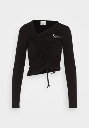 Fekete, hosszú ujjú felső V-nyakkal és elöl ráncolt részletekkel. Állítható zsinórok találhatók az alján, valamint egy fehér logó.