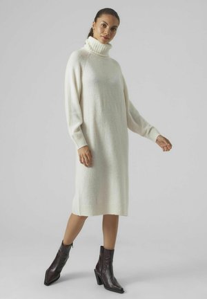 Kleid aus cremefarbenem Strick mit hohem, geripptem Rollkragen, langen Ärmeln und gerader Silhouette, kombiniert mit dunkelbraunen Stiefeletten.