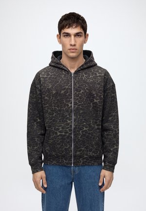 Jeune homme aux cheveux courts et sombres portant un sweat à capuche zippé gris foncé imprimé léopard et un jean bleu, debout devant un fond uni.