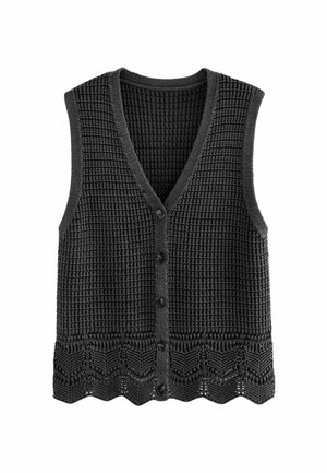 Femme portant un gilet noir en maille sans manches boutonné et un pantalon taille haute imprimé léopard, les mains dans les poches, sur un fond uni.