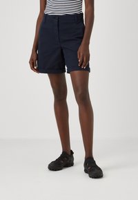 Pantaloni corti blu navy su misura con risvolti, realizzati in un tessuto morbido. Abbinati a scarpe sportive nere. Presentano una chiusura con bottone frontale.
