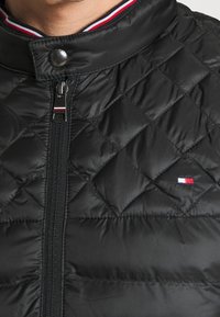 Prošívaná černá bunda s vysokým límcem, má přední zip a zapínání na patentky, zdobená tříbarevným pruhem na límci.
