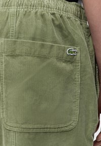 Gros plan sur la poche d'un pantalon en velours côtelé vert olive avec un logo crocodile blanc et vert en haut à droite.
