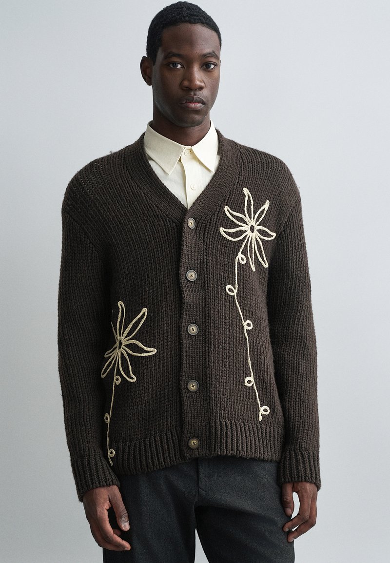 Barna kötött kardigán V-nyakú fazonban, elején gyöngyvirág motívumú krémszínű hímzéssel, gombos záródással, hosszú ujakkal, bordázott mandzsettákkal és aljjal.