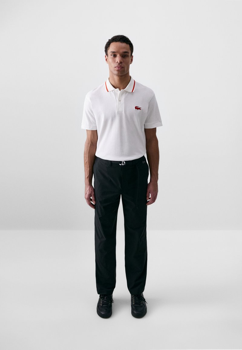 Chemise polo blanche avec bordure de col rouge et logo Lacoste, associée à un pantalon noir ample et des baskets noires. Tenue simple et décontractée.