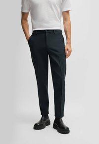 Dark tailored trousers with a slim fit, featuring a smooth fabric, straight legs, and a single button closure. Paired with black boots.